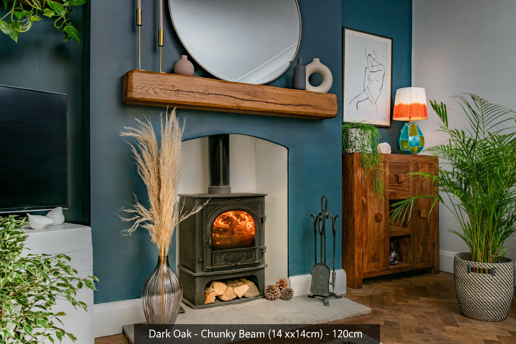 A dark oak fireplace beam mounted on a blue wall above a fireplace, with decorative mirrors and lighting fixtures, surrounded by indoor plants and a side cabinet.