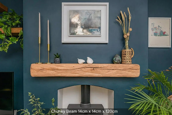 A natural oak fireplace beam with visible knots and splits, placed on a fireplace mantle against a blue wall, with decorations and plants around it.